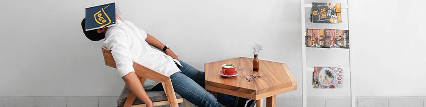 Person resting head on table with a book covering face in a minimalist cafe setting